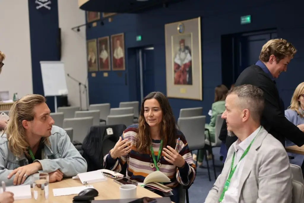 A group of people sit around a table engaged in discussion. A woman in the center gestures while talking, with notebooks and coffee cups on the table. Portraits hang on the dark blue wall in the background.