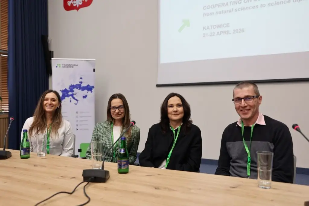Four people sit at a conference table with microphones and water bottles. Behind them is a screen displaying event details about a science cooperation conference in Katowice, April 2026.