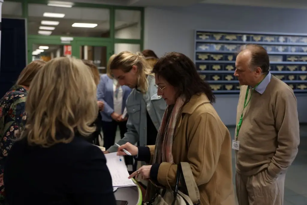 A group of people stands at a registration table indoors. Some are signing papers while others wait. One man wears a lanyard and name tag. The background shows green doors and a wall with display shelves.