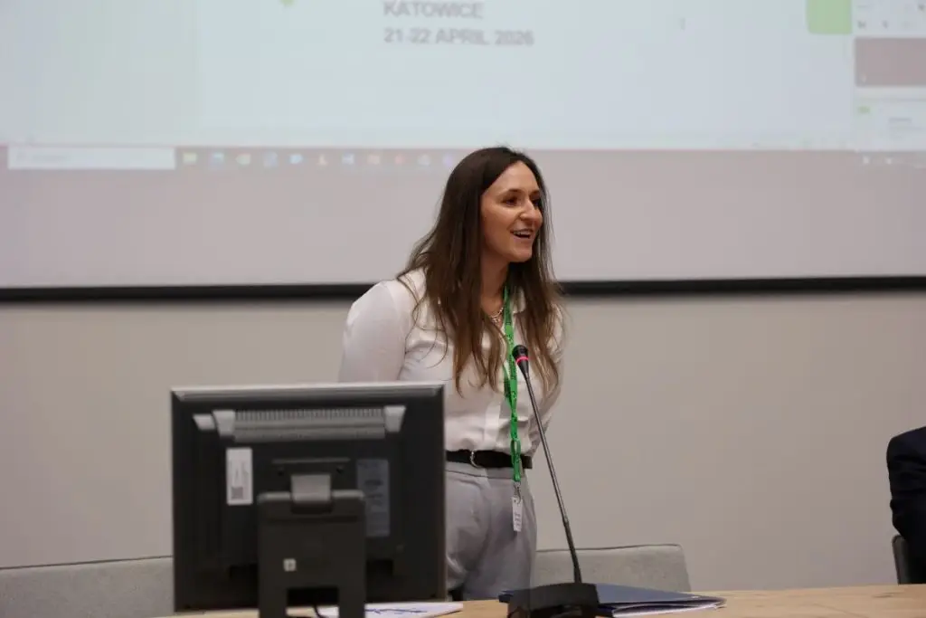 A woman with long brown hair stands and speaks at a podium with a microphone, in front of a projection screen displaying event details. She is wearing a white blouse and gray pants. A computer monitor is on the desk in front of her.