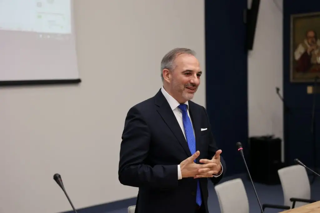 A man in a suit and blue tie speaks while standing in a conference room with microphones and empty chairs. A projection screen and a framed portrait are visible in the background.