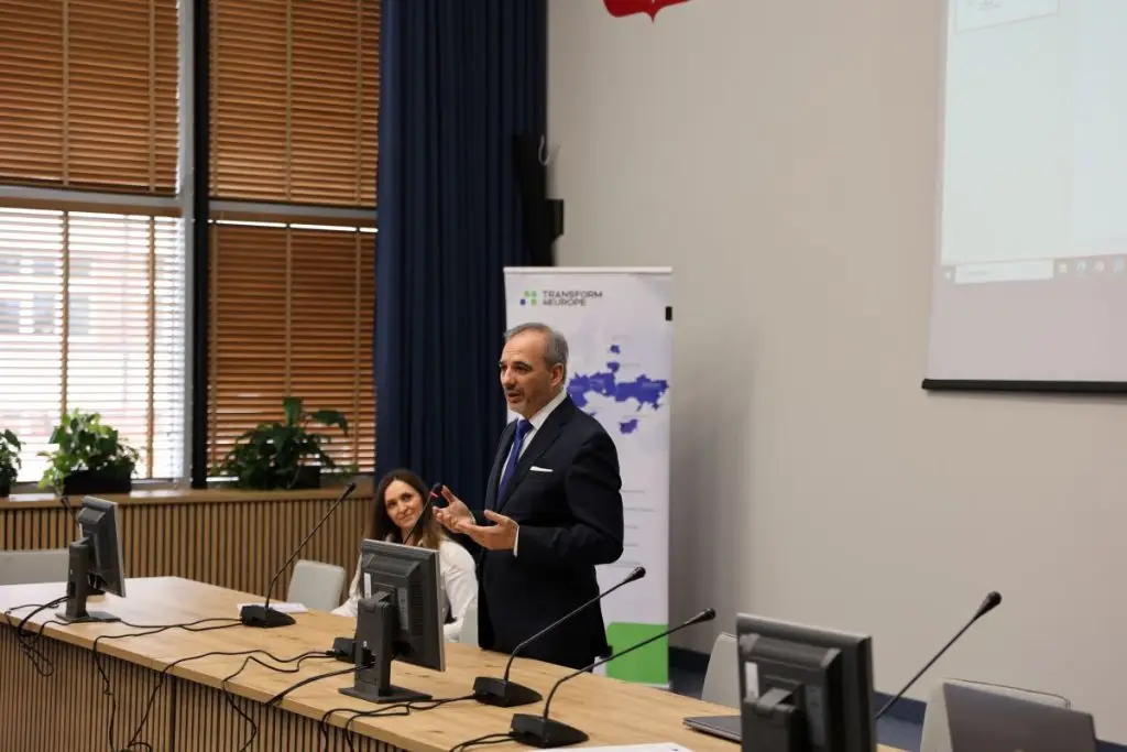 A man in a suit is speaking and gesturing at the front of a conference room, while a seated woman listens. Several monitors and microphones are on the table, and a presentation banner is in the background.
