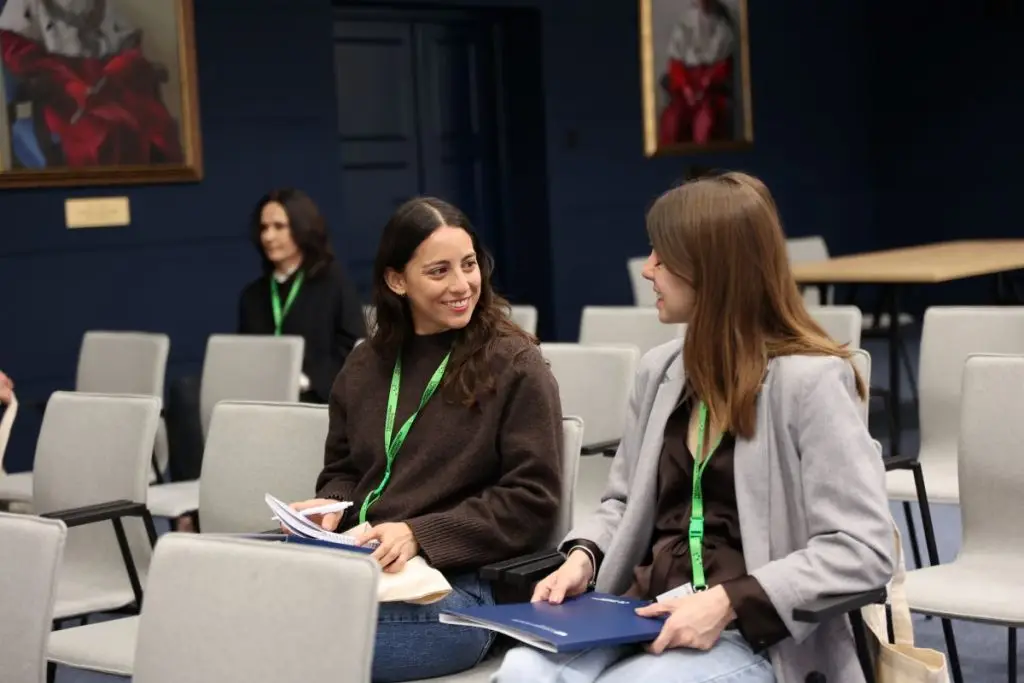 Two women with green lanyards sit and smile at each other in a conference room with empty chairs. A woman sits further back, and paintings are visible on the dark blue walls in the background.