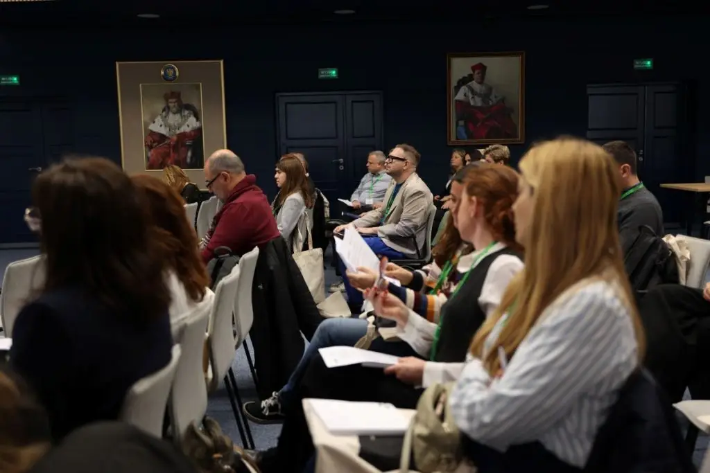 People seated in rows at an indoor conference or seminar, listening attentively and holding papers. Two large framed portraits hang on the dark wall in the background.