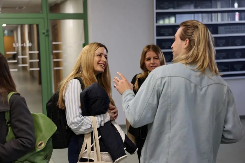 Three young adults stand and talk in a hallway, smiling and engaged in conversation. Two women face a man with long blond hair. One woman holds a bag and jacket. A green door and notice board are visible in the background.