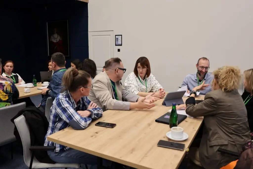 A group of people sit around two tables in a conference room, engaged in conversation. Some have laptops open, and there are cups, bottles, and notebooks on the tables. The atmosphere appears collaborative and focused.