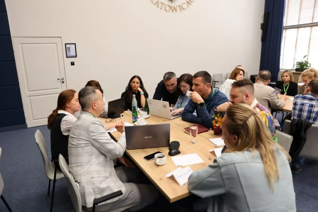 A group of people sit around a table in a meeting room, engaged in discussion with laptops, papers, and drinks on the table. Some are talking, while others are listening attentively.