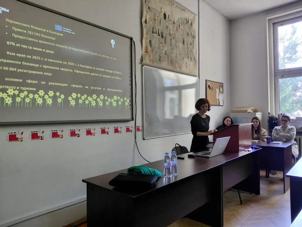 A woman stands at a lectern presenting a slideshow to three seated people in a classroom. The slide projects information and graphics in Bulgarian on a wall. Sunlight enters through large windows on the right.