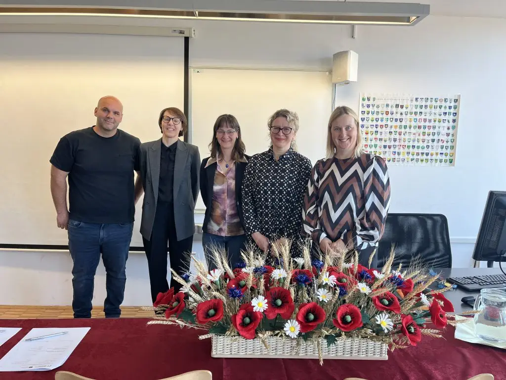 Five people stand together in a classroom behind a table decorated with a basket of red, white, and blue artificial flowers. There are whiteboards and a colorful chart on the wall behind them.