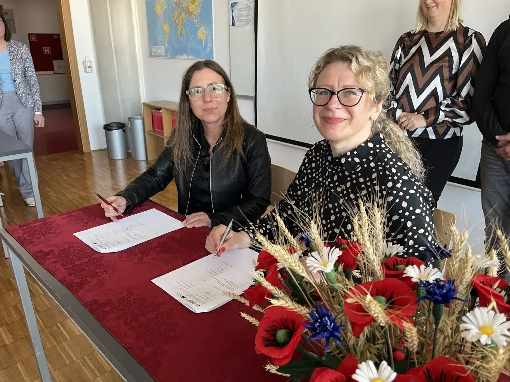 Two women sit at a table with red cloth, signing documents and smiling at the camera. A bouquet of red, white, and blue flowers is on the table. Other people stand in the background in a bright room.