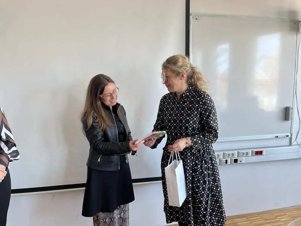 Two women stand in front of a whiteboard; one woman smiles as she hands a book and envelope to the other, who holds a white gift bag. Both are dressed professionally and appear to be engaged in a friendly exchange.
