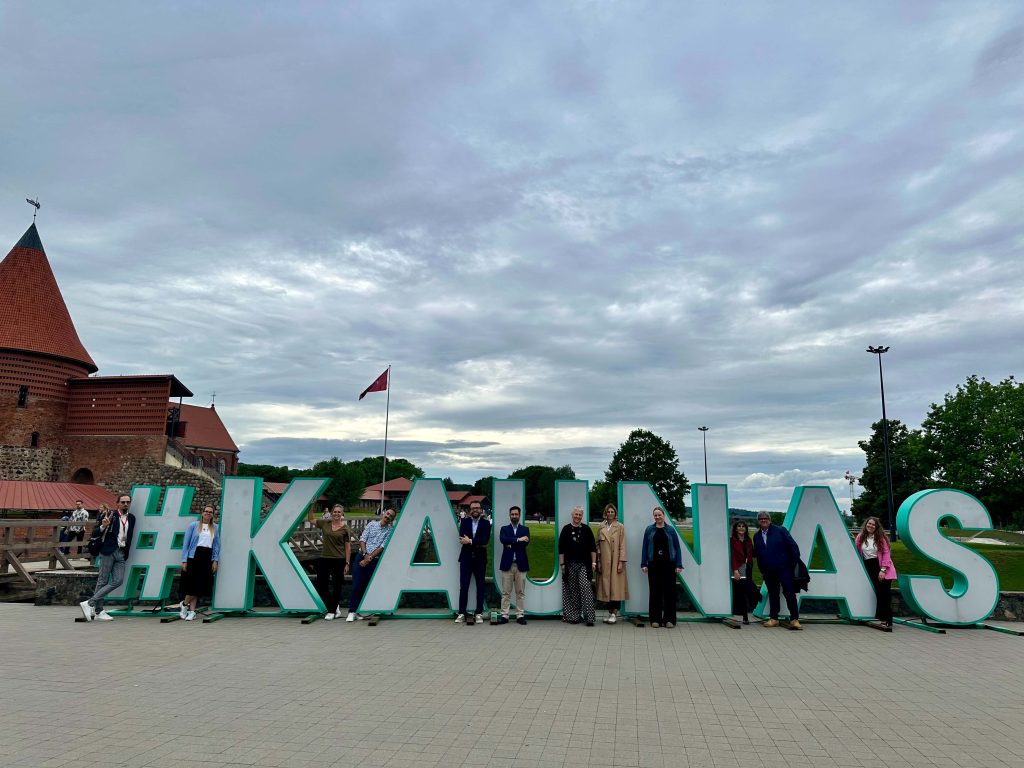 A group of people stand in front of large green and white letters spelling #KAUNAS outdoors, with a historic brick building and cloudy sky in the background.