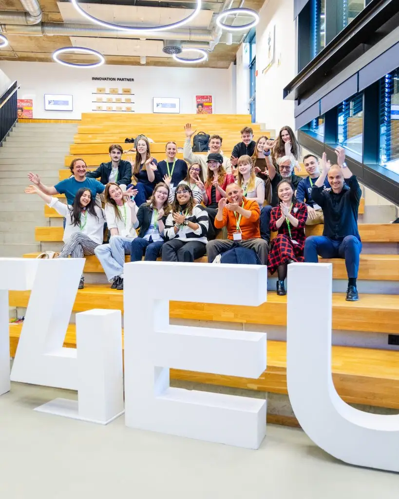 A group of people sit and stand on wooden bleachers, smiling and waving at the camera. Large white letters in the foreground spell “4EU.” Modern lights and a staircase are visible in the background.