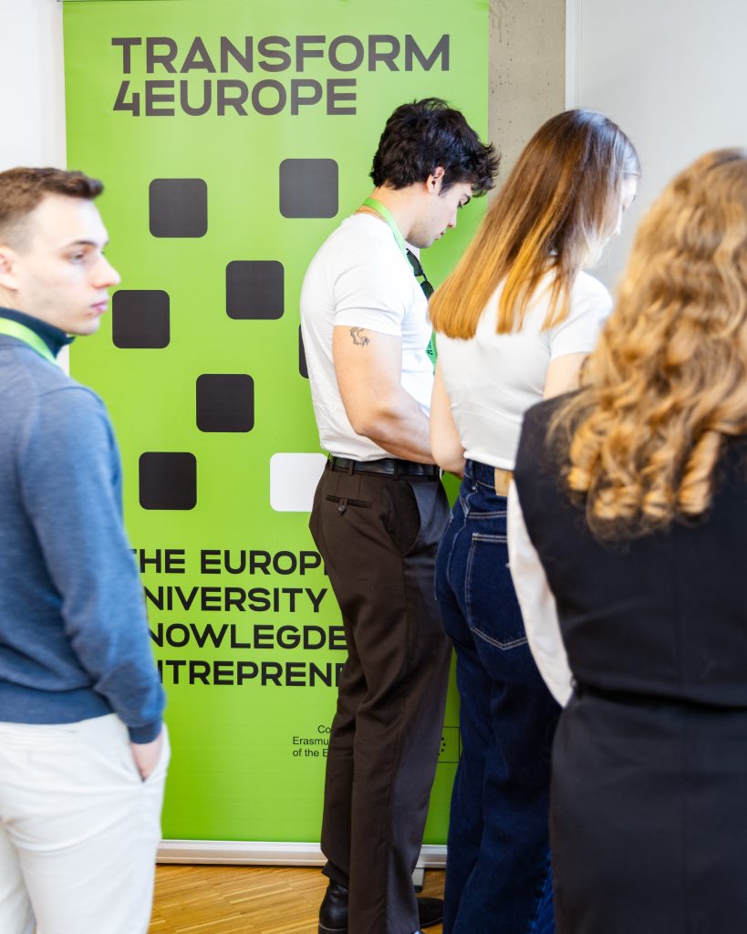 Four people stand in front of a bright green Transform 4 Europe banner at an event. Three of them are facing away, and one looks to the side. The banner promotes knowledge and entrepreneurship.