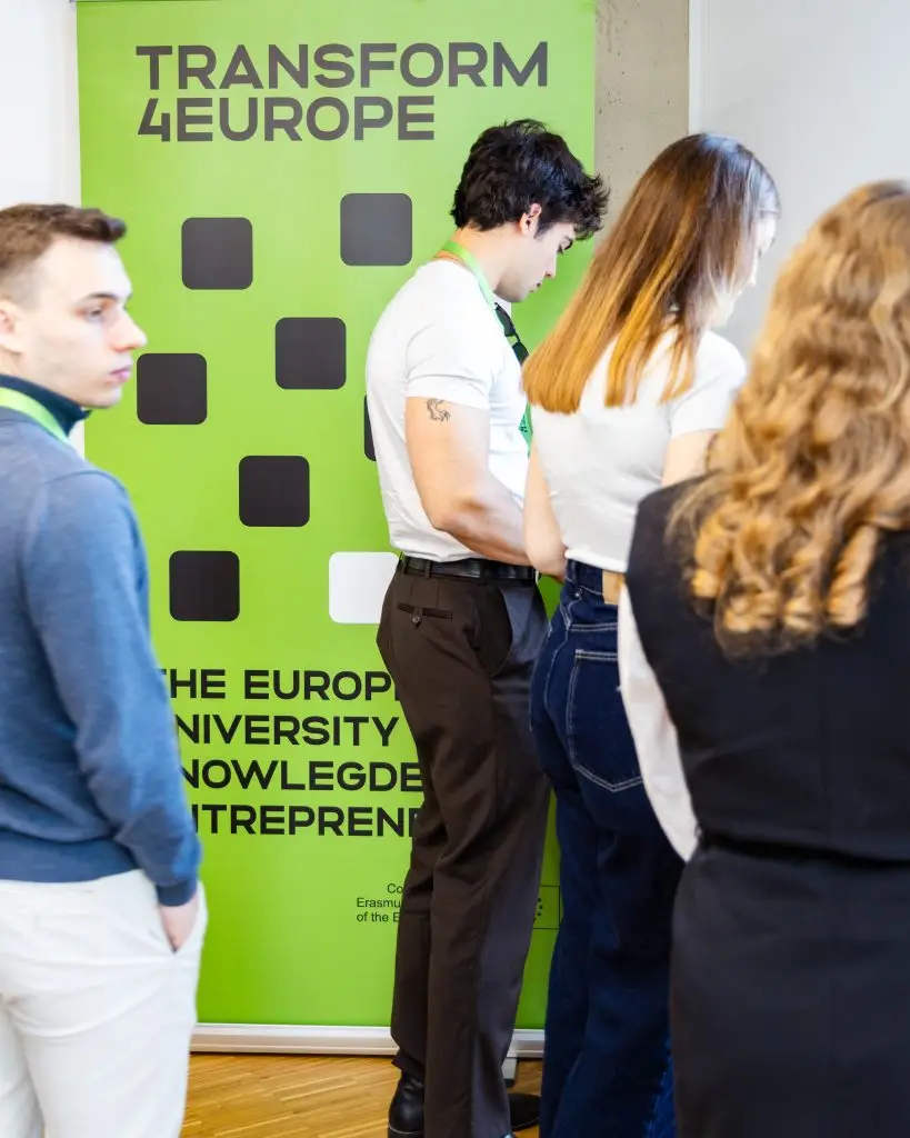 Four people stand in front of a bright green Transform 4 Europe banner at an event. Three of them are facing away, and one looks to the side. The banner promotes knowledge and entrepreneurship.
