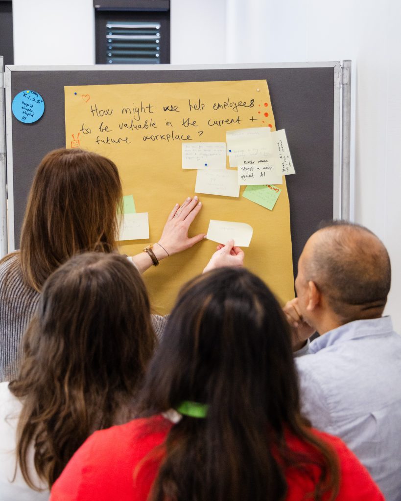 Four people are gathered around a board with sticky notes, collaborating and discussing ideas under the handwritten question, “How might we help employees be valuable in the current and future workplace?”.