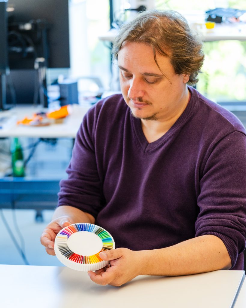 A person in a purple sweater sits at a desk holding a circular color wheel with various colored sections, looking down at it thoughtfully. Office equipment and a window are visible in the background.