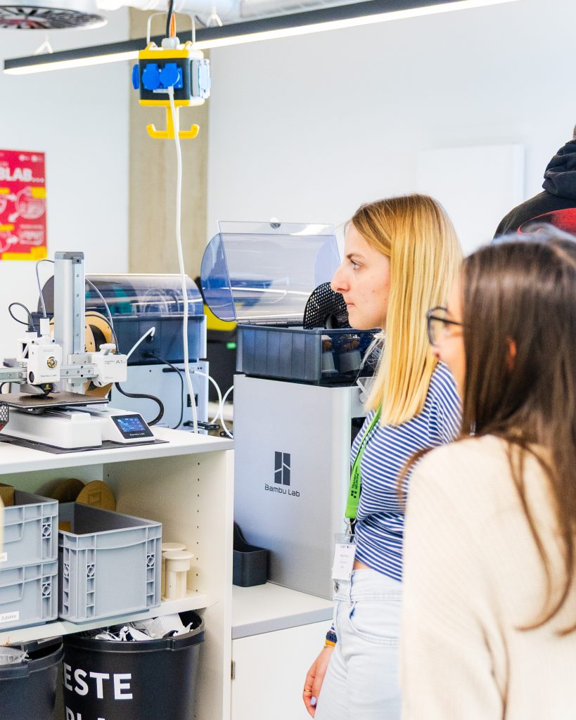 Two women observe a 3D printer at work in a brightly lit lab, surrounded by equipment, plastic bins, and technology. One woman wears a striped shirt and the other has glasses.
