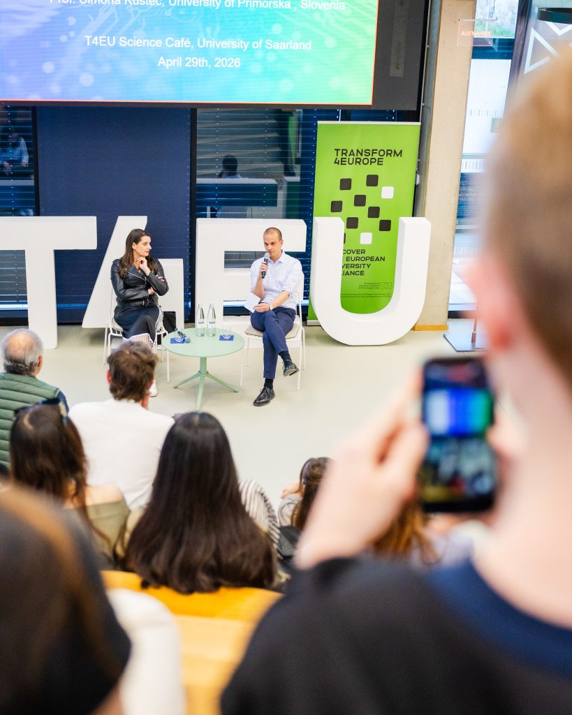 Two people sit on a stage in front of large white T4EU letters, speaking to an audience. A person in the foreground is taking a photo with a smartphone. A green Transform Europe banner is visible behind the speakers.