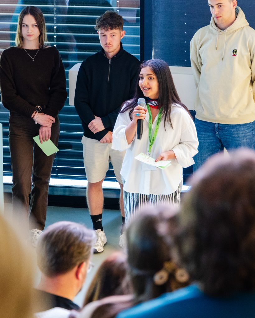 A young woman holding a microphone speaks to an audience, while three young adults stand behind her, listening attentively. The scene appears to be a presentation or group discussion indoors.