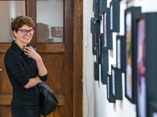 A woman with short hair and glasses smiles while viewing framed artwork on a gallery wall. She is wearing a black dress and holding a bag in one hand.
