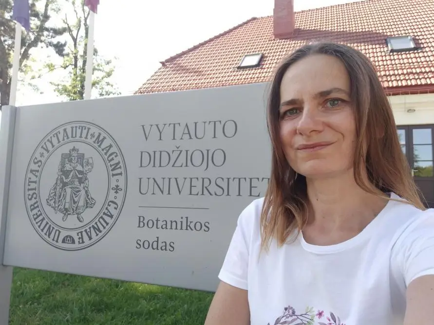 A woman with straight brown hair takes a selfie in front of a sign for Vytauto Didžiojo Universitetas Botanikos sodas, with a building featuring a red-tiled roof in the background.