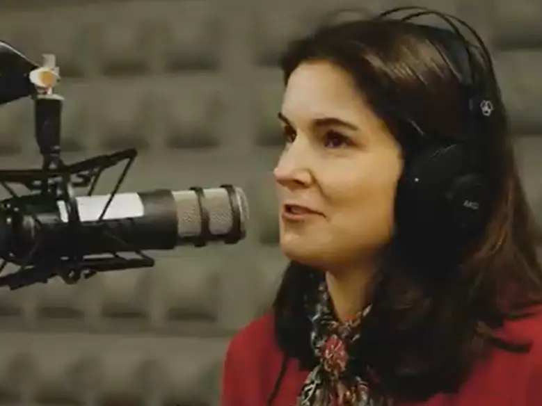 A woman with brown hair wearing headphones speaks into a microphone in a recording studio with soundproof foam panels on the walls.