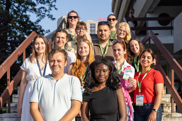 A group of thirteen people, smiling and standing together on outdoor steps in the sunlight, wearing lanyards and casual summer clothing. Trees and buildings are visible in the background.