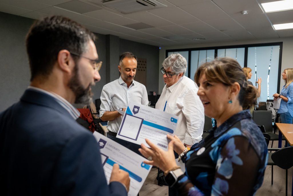 Four adults in business attire talk and hold certificates in an office meeting room, with other people conversing in the background. The atmosphere appears casual and professional.