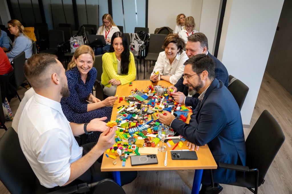 Six adults in business attire sit around a table covered with LEGO bricks, smiling and engaging in a group activity. Other people are visible working in the background. The setting appears to be an office or meeting room.