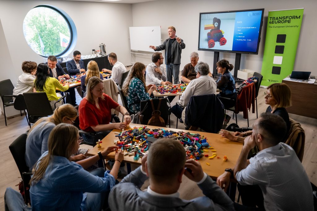 A group of adults sit around tables covered with colorful LEGO pieces, actively building and discussing, while a presenter stands by a screen displaying a LEGO model in a modern meeting room.