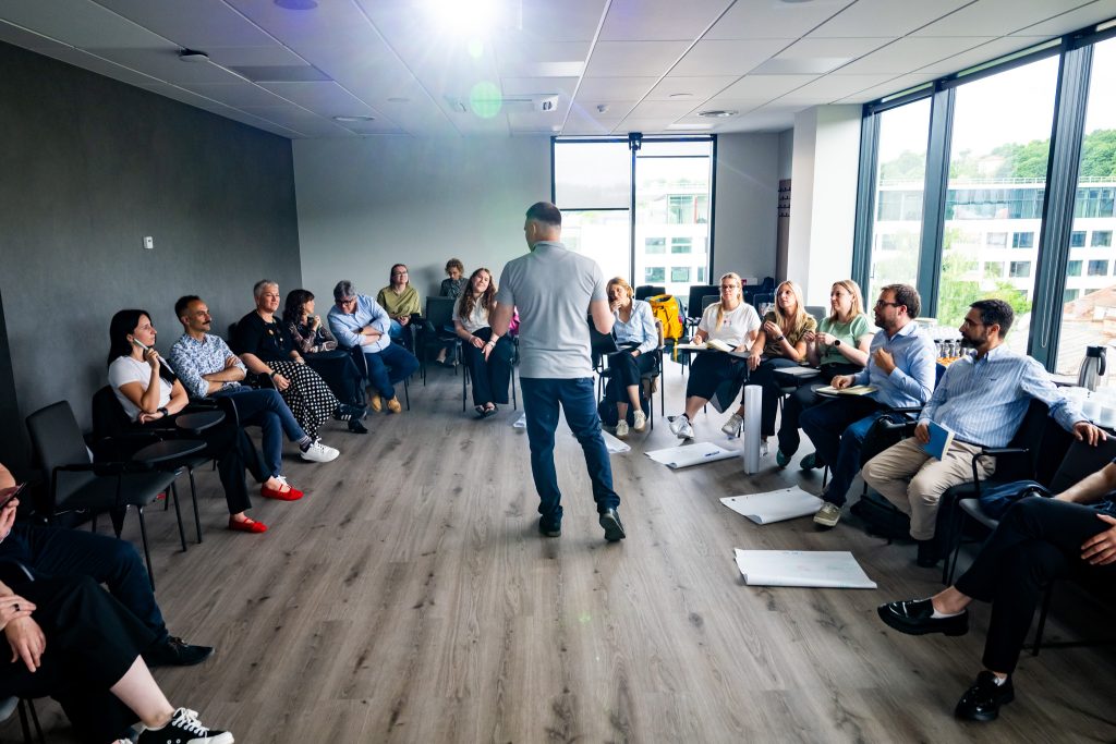 A man stands in the center of a modern, bright room speaking to a seated group of adults arranged in a circle, suggesting a workshop or group discussion. Papers and notebooks are scattered on the floor and tables.