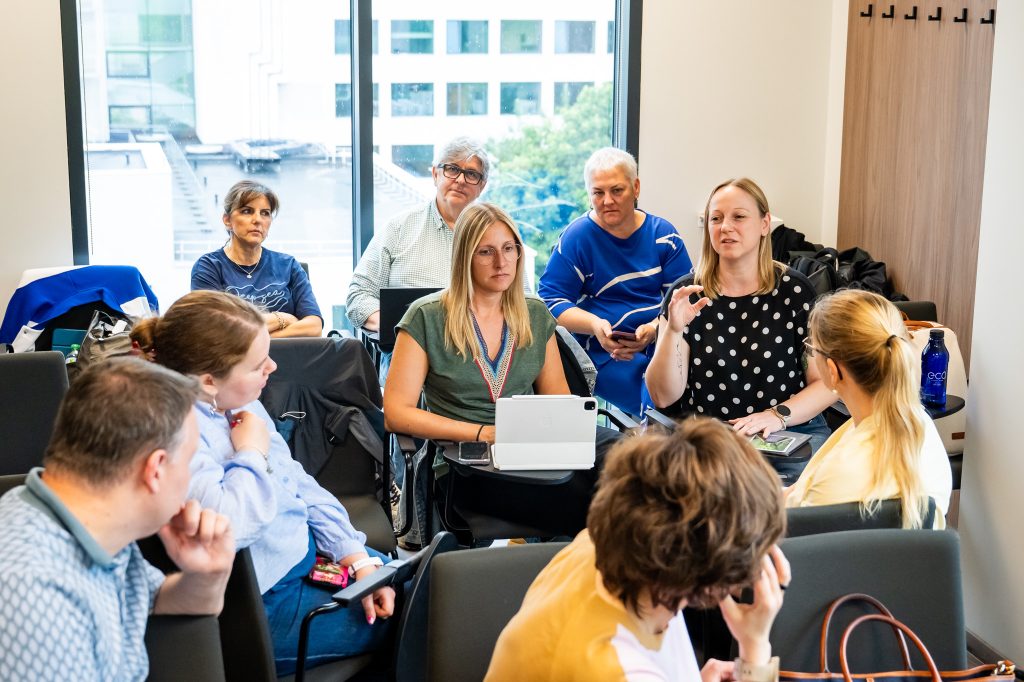 A group of people sits in a modern office, engaged in a discussion. Some are listening while others are speaking. There is a large window in the background with a view of buildings and trees outside.