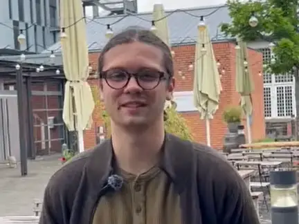 A young person with long hair tied back, wearing glasses and a brown jacket, stands and smiles in an outdoor café area with closed umbrellas, string lights, and brick buildings in the background.