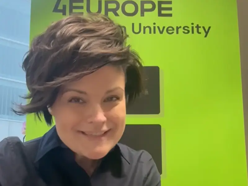 A person with short dark hair and a black shirt smiles in front of a bright green sign that reads 4EUROPE and University in black text.