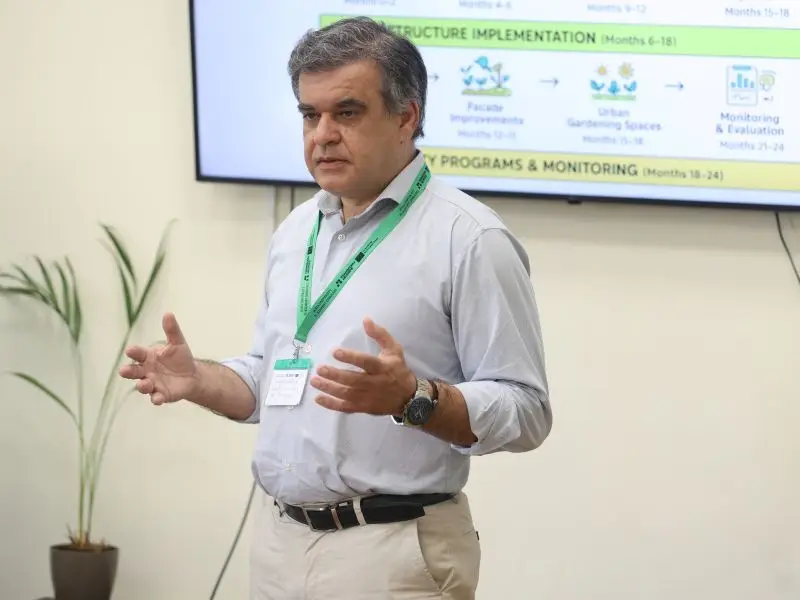 A man wearing a green lanyard and name badge gestures while speaking in front of a projection screen with charts and text. A plant is visible in the background.