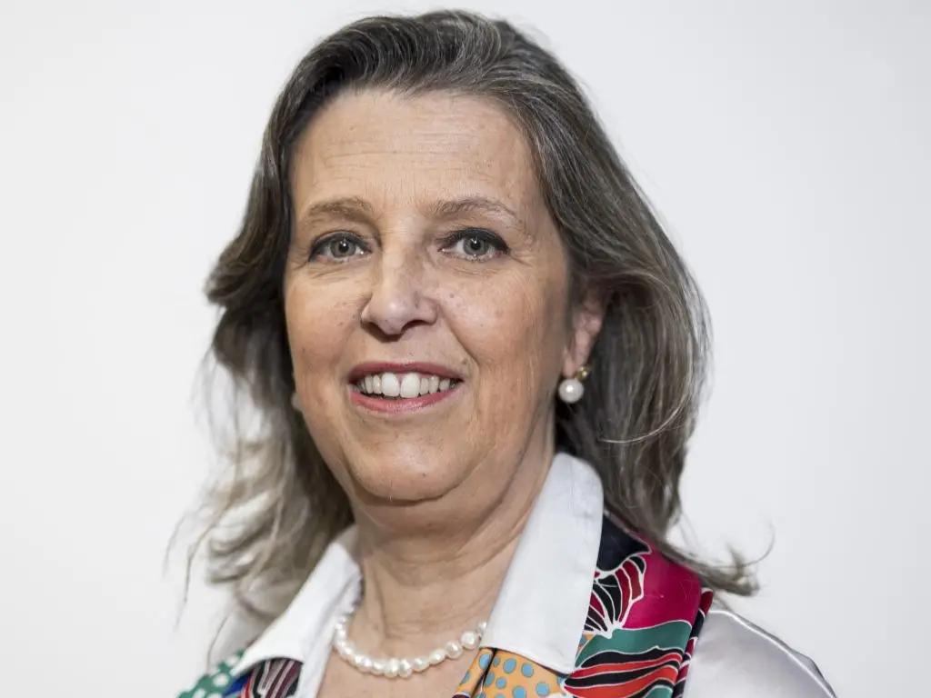 Smiling middle-aged woman with gray hair wearing a colorful scarf, a white blouse, pearl earrings, and a pearl necklace, posing against a plain white background.