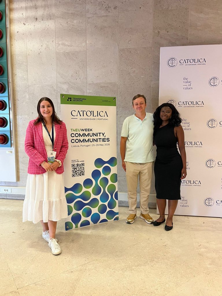Three people stand indoors beside a green Católica University poster and a branded backdrop. The woman on the left wears a white dress and pink jacket; the man in the center wears a white shirt; the woman on the right wears a black dress.