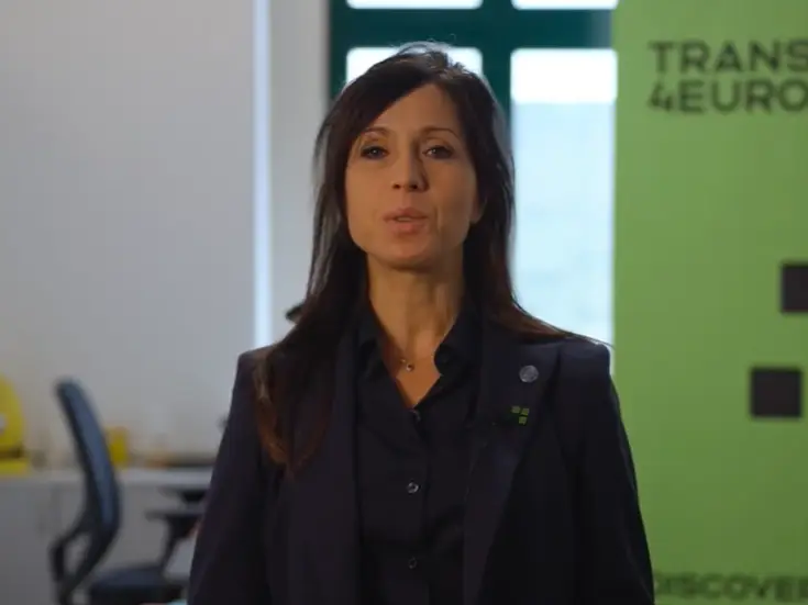 A woman with long dark hair, wearing a dark blazer and blouse, stands indoors in front of a green banner with partial text reading Transform4Europe. An office setting with chairs and a window is in the background.