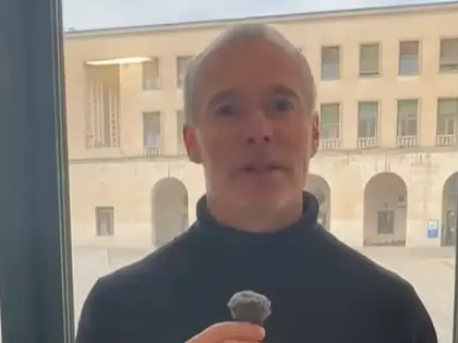A man with short gray hair wearing a black turtleneck holds a small microphone while standing indoors, with a courtyard and a beige building visible through the window behind him.