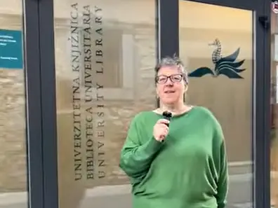 A person wearing glasses and a green sweater stands in front of glass doors with University Library and its translation written on them, holding a small microphone. A seahorse logo is also visible on the door.