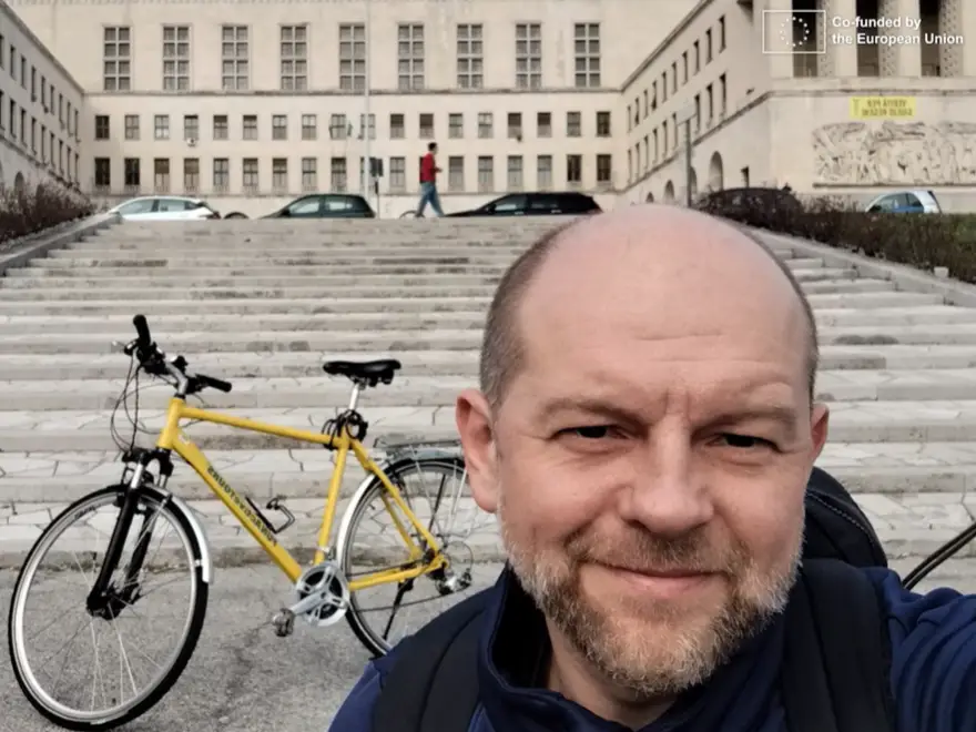 A man smiles for a selfie in front of a large building with steps. A yellow bicycle is parked nearby, and a person walks in the background. A logo in the top right corner reads Co-funded by the European Union.
