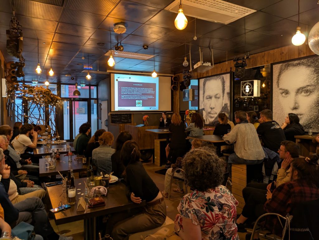 A group of people sit at tables in a cozy, dimly lit café watching a presentation on a screen. Large black-and-white portraits decorate the walls, and warm hanging lights illuminate the space.