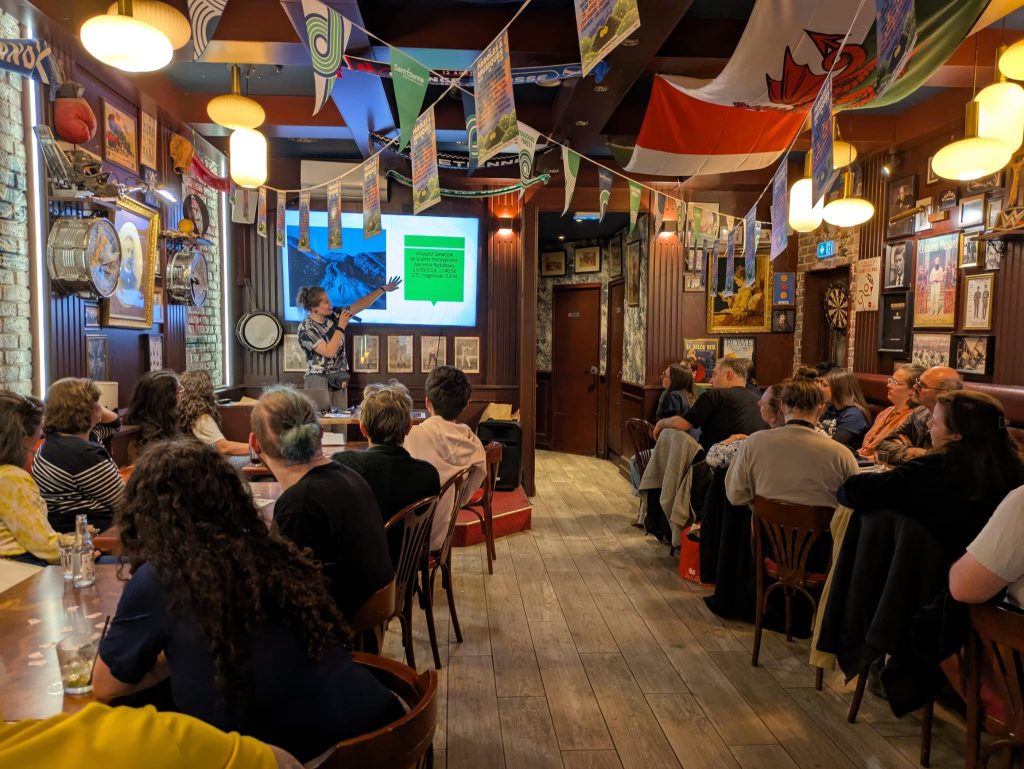 A group of people sit at tables in a cozy pub, attentively watching a presenter who is pointing at a green slide on a screen. Flags and decorations hang from the ceiling, and framed pictures line the walls.