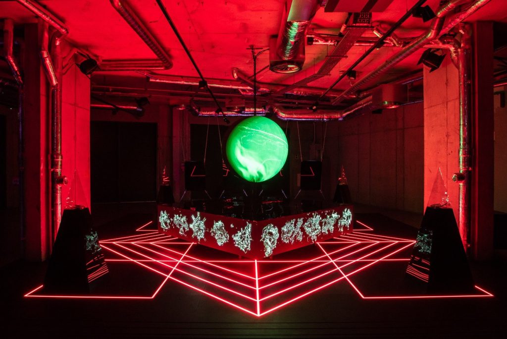 A futuristic art installation with red neon lights forming geometric patterns on the floor, a large glowing green orb suspended above a platform, and exposed pipes on the ceiling in an industrial setting.