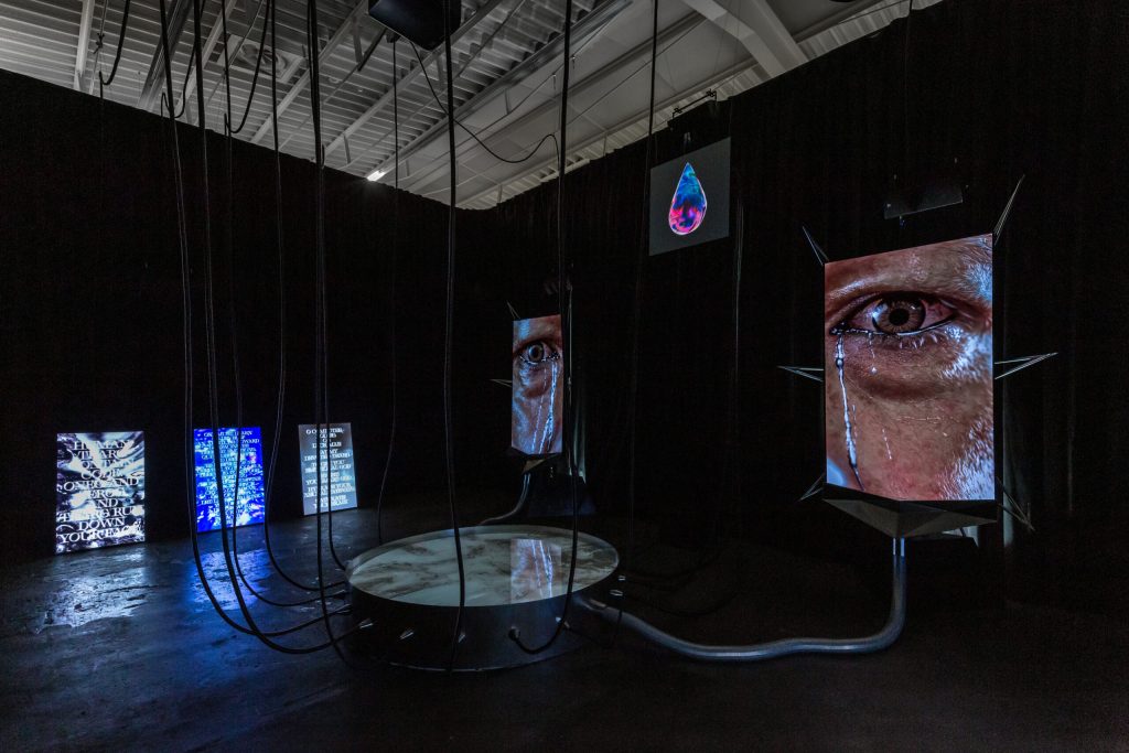 A dark room with large digital screens showing a close-up of a human eye and abstract visuals; cables hang from the ceiling, and a round mirror or platform is on the floor, creating a futuristic, immersive installation.