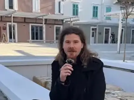 A person with long hair and a beard, dressed in a black coat, holds a microphone while sitting on a white bench in an outdoor urban area with modern buildings in the background.