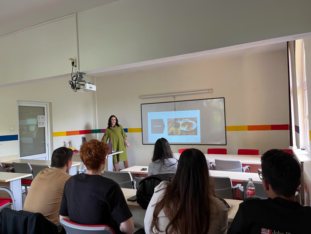 A person stands at the front of a classroom giving a presentation, pointing at a screen displaying an image of food. Several students sit at desks facing the presenter.
