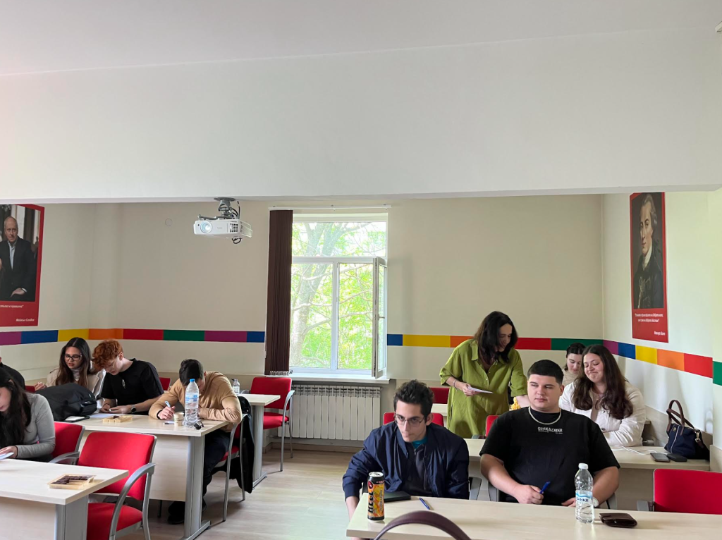A classroom with students seated at desks, some working in pairs and others alone. Two large portraits hang on the walls, and a window lets in natural light. The atmosphere appears relaxed and collaborative.