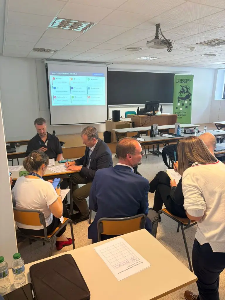 A group of people in business attire sit and stand around tables in a classroom, engaged in discussion. A projection screen with charts and a green event banner are visible in the background.